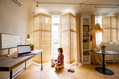 Young woman meditates after work, sitting on the floor at cozy home office. Stylish interior with workplace and computers in front. Mental health and relaxation, working from home concept