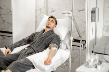 Man in bathrobe lying on medical couch during blood wash procedure with catheter in his arm at medical centre