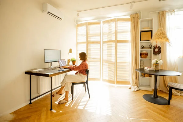 自宅の日当たりの良いリビングルームで居心地の良い職場に座っている間 女性はコンピュータ上で動作します ホームオフィスからのリモートワークの概念 — ストック写真