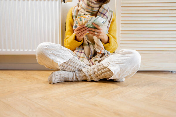 Warmly dressed woman counting money while sitting near radiator at home. Concept of expensive energy resources and the energy crisis in Europe
