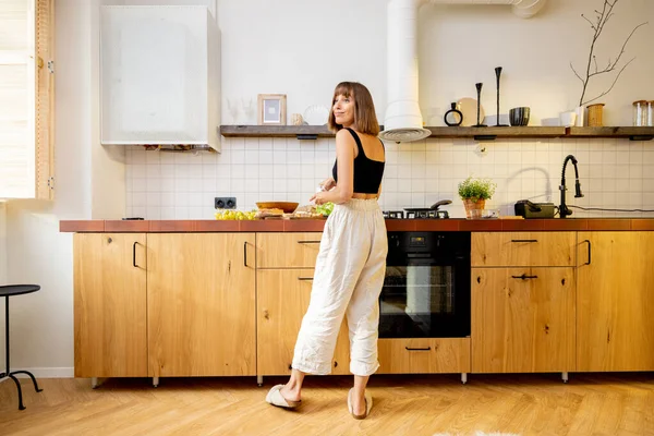 一个年轻快乐的女人站在厨房桌旁 在家里吃着健康的素食的画像 现代舒适的厨房内部视野开阔 — 图库照片