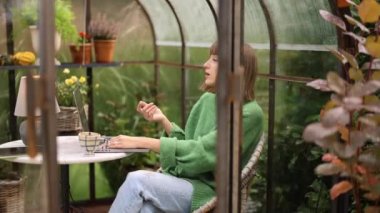 Genç kadın dizüstü bilgisayarda çalışıyor, arka bahçesinde çiçekler ve bitkilerle birlikte cam evde yuvarlak masa başında otururken internetten konuşuyor. Doğa üzerine rahat bir atmosferde evden çalışmak