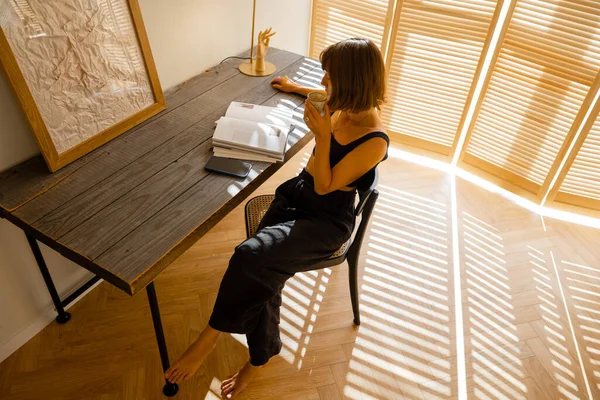 Woman reads some magazine while sitting at cozy workplace by the wooden table at home. Living room in beige tones with window blinds on background