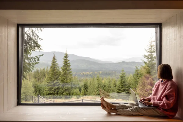 Woman works on laptop while sitting on a window with great view on mountains. Remote work from cozy place and escaping to nature concept