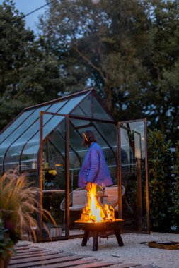 Kadın boş zamanlarını arka bahçede akşam pikniğinde şenlik ateşiyle geçirir. Arka planda güzel cam turuncu ev
