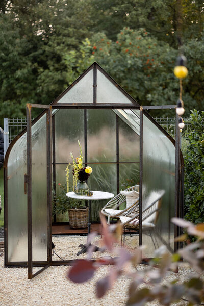 Beautiful vintage greenhouse made of rusty metal and glass with table and flowers inside at backyard
