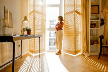 Young woman stands alone by the window blinds at cozy and sunny living room of modern apartment in beige tones. Quarantine, loneliness and life at home