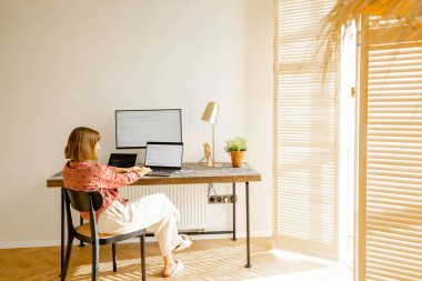 Woman works on computers while sitting by a cozy workplace in sunny living room at home. View from the backside. Concept of remote work from home office. Mockup image