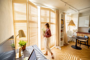Woman walks with a vase of flowers, decoration and having some house chores at home. Wide interior view on sunny studio apartment in beige tones