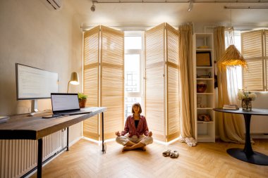 Young woman meditates after work, sitting on the floor at cozy home office. Stylish interior with workplace and computers in front. Mental health and relaxation, working from home concept