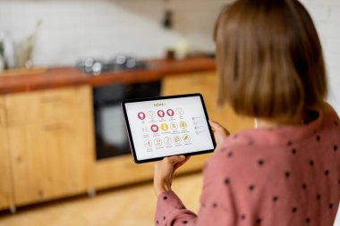 Woman holds a digital tablet with running mobile application for smart home, standing in kitchen of modern apartment. Controling smart devices with a digital tablet