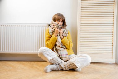 Sıcak giyimli kadın, ısıtmanın yüksek fiyatına üzülüyor. Evdeki akünün yanında üşüyor.