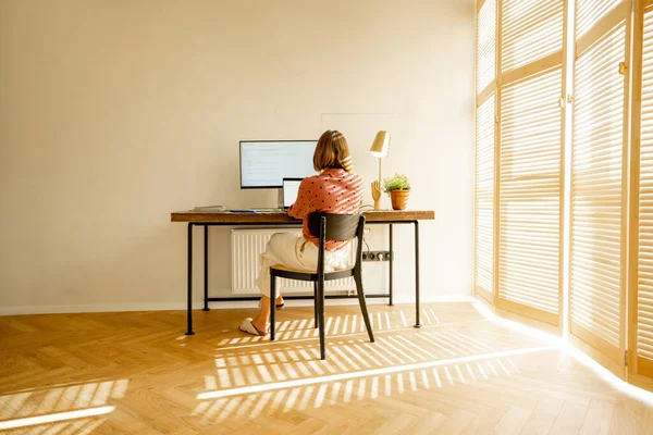 窓にブラインドがある日当たりの良いリビングルームで居心地の良い職場に座っている間 女性はコンピュータ上で動作します ホームオフィスからのリモートワークの概念 — ストック写真