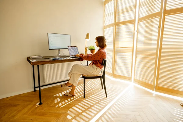 Woman works on computers while sitting by a cozy workplace at sunny living room with blinds on the window. Concept of remote work from home office