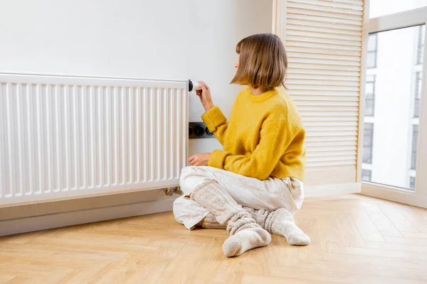 Woman controls room temperature with a thermostat on radiator, feeling cold at home. Concept of saving energy resources and the energy crisis