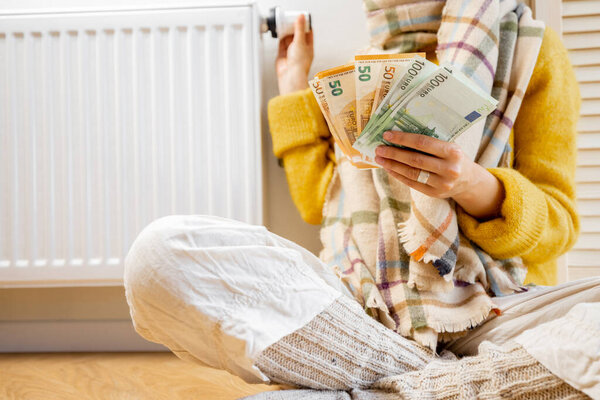 Warmly dressed woman counting money while sitting near radiator at home. Concept of expensive energy resources and the energy crisis in Europe