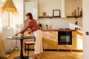 Kadın yemek masasına çiçek koyar, dekorasyon yapar ve evde ev işleri yapar. Stüdyo dairesinin içi bej renklerle güneşli.