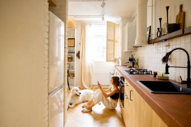 Woman uses smart phone while sitting with her dog in kitchen. Interior view on modern and stylish kitchen in beige tones. Domestic lifestyle concept