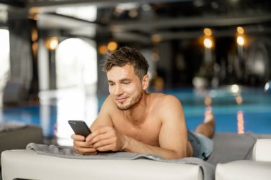 Man rests by the swimming pool indoors, talking on phone while lying relaxed on lounge bed indoors. Man spends holidays at spa resort with an indoor pool