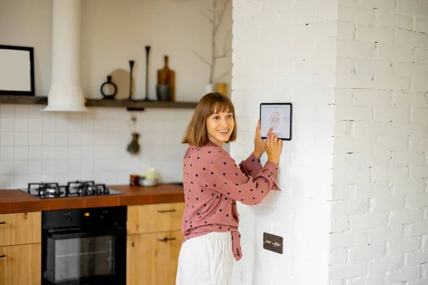 Woman controls room temperature on a digital panel mounted on the wall in kitchen. Concept of smart home and new technologies for home comfort