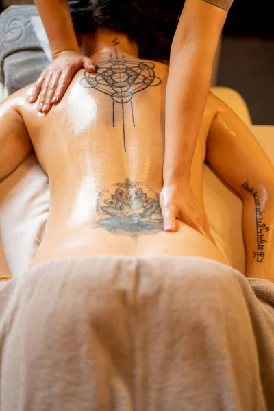 Woman receiving professional relaxing back massage at Spa salon. Client lying covered with towel on medical couch with tattoos on her back