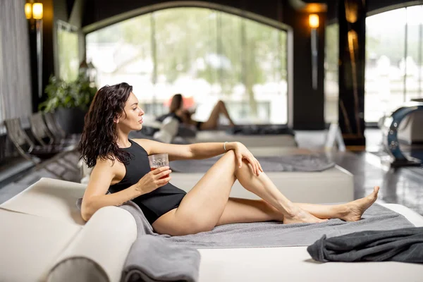 Adult woman rests by the swimming pool indoors, drinking while lying relaxed on lounge bed indoors. Woman spends holidays at spa resort with an indoor pool