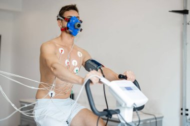 Man athlete with breath mask and electrodes training on bike simulator, examining his cardiovascular system at medical office