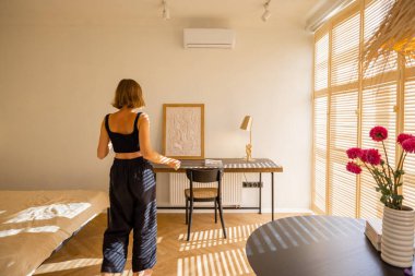 Güneşli stüdyo dairesinin içinde bej renkte bir daire, çalışma yeri ve evde boş zaman geçiren bir kadın.