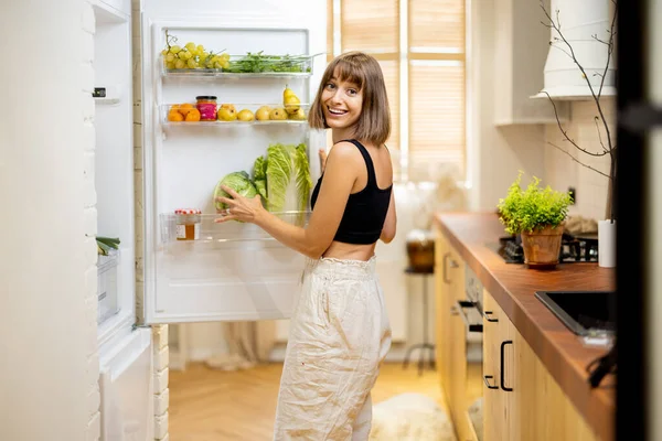 年轻女人在家里做饭的时候从冰箱里取些吃的 冰箱里塞满了健康的蔬菜和水果 健康生活方式和饮食的概念 — 图库照片