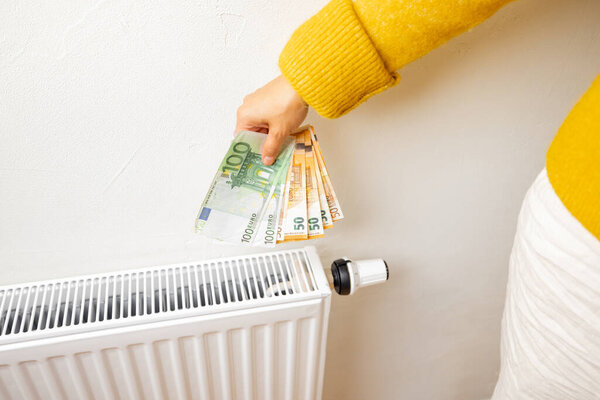 Person holding euro banknotes near heater at home. Concept of expensive energy resources and the energy crisis in Europe
