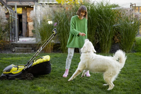 若い女性は彼女の白い愛らしい犬と遊ぶ 彼女のコテージの裏庭でペットと余暇時間を過ごす 芝刈り機でガラスを切る — ストック写真