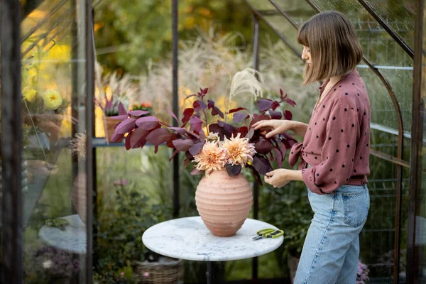 Woman florist makes a beautiful and lush bouquet in vase on a table in a small greenhouse for plants at back yard. Floristry and beauty concept