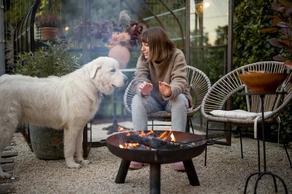 若い女性は手を暖め 夜を過ごす彼女の犬によって火で居心地の良い美しい裏庭で — ストック写真