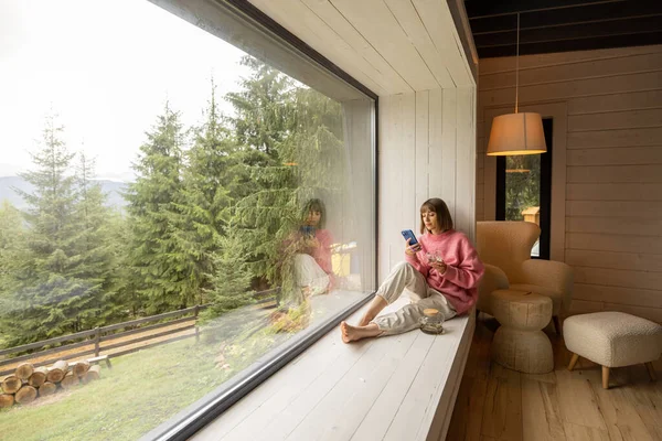 Kadın Pencere Pervazında Telefonla Oturur Doğadaki Ahşap Evde Dinlenirken Dağların — Stok fotoğraf
