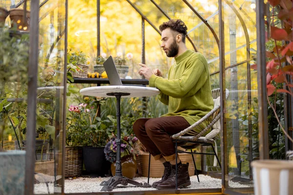 裏庭の植物や花とガラスの家の丸いテーブルに座っている間 男はノートパソコンで動作します 自然の中で居心地の良い雰囲気で自宅から仕事 — ストック写真