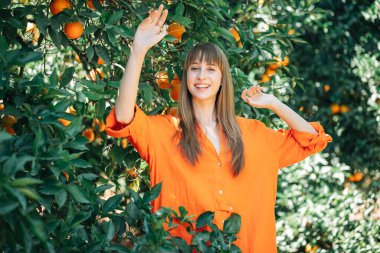 Turuncu elbiseli mutlu kız elini kaldırıp portakal bahçesine bakıyor. 