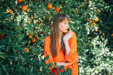 Turuncu elbiseli genç kız turuncu bahçedeki sağ elini kaldırarak sağına bakıyor. 