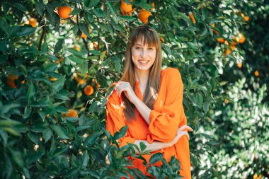 Turuncu elbiseli mutlu kız turuncu bahçede kameraya poz veriyor. 