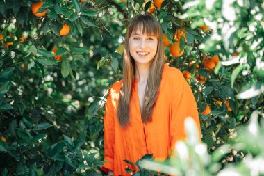 Turuncu elbiseli gülümseyen genç kız turuncu bahçedeki kameraya bakıyor. 