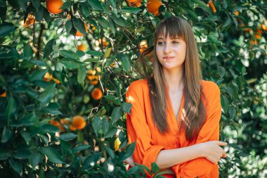 Turuncu elbiseli mutlu kız, turuncu bahçede kollarını kavuşturarak başka tarafa bakıyor. 