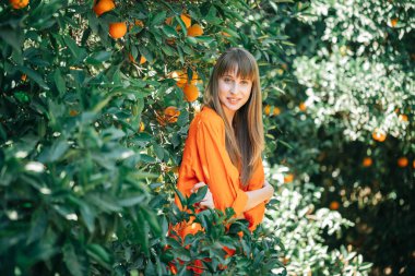 Turuncu elbiseli gülümseyen kız turuncu bahçede kollarını kavuşturarak kameraya bakıyor. 