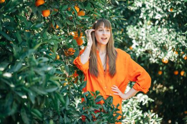 Turuncu elbiseli mutlu kız elini portakal bahçesinde beline koyarak yüzünü çeviriyor. 