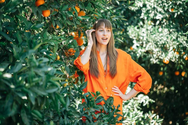 Turuncu elbiseli mutlu kız elini portakal bahçesinde beline koyarak yüzünü çeviriyor. 