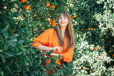 Turuncu elbiseli mutlu genç kız turuncu bahçede gülümseyerek kameraya bakıyor. 