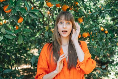 Turuncu elbiseli genç kız ellerini portakal bahçesinde tutarak kameraya poz veriyor. 