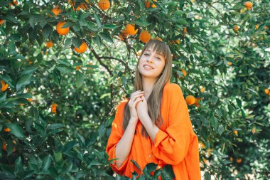 Turuncu elbiseli mutlu genç kız, portakal bahçesinde el ele tutuşarak kameraya bakıyor. 