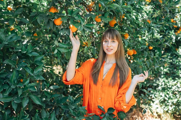 Turuncu elbiseli mutlu kız ellerini portakal bahçesinde kaldırarak kameraya poz veriyor. 