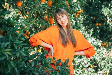 Turuncu elbiseli gülümseyen genç kız elini portakal bahçesinde beline koyarak kameraya poz veriyor. 