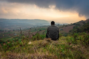 Dağın tepesinde oturan genç adam sisli tepe arkaplanı ve sabah dramatik gökyüzü ile Badda Peak Shillong Meghalaya Hindistan 'da çekilmiştir..