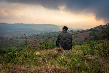 Dağın tepesinde oturan genç adam sisli tepe arkaplanı ve sabah dramatik gökyüzü ile Badda Peak Shillong Meghalaya Hindistan 'da çekilmiştir..